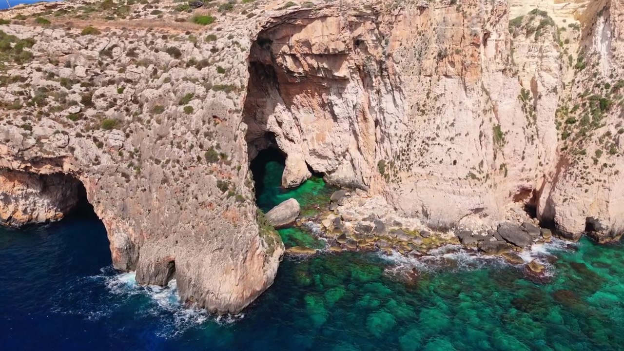 Maltas Blaue Grotte, ein majestätischer natürlicher Kalksteinbogen und Höhlenkomplex am Meer an der Südküste. Weitläufige Luftaufnahmen zeigen tief azurblaues Wasser, das mit schroffen, sonnenverwöhnten Klippen kontrastiert.