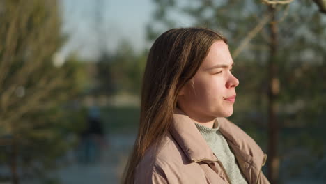 close-up of a girl carrying a skateboard while walking through a park on a sunny day. she is wearing a peach jacket and a grey inner shirt, with her mouth closed, enjoying the calm environment