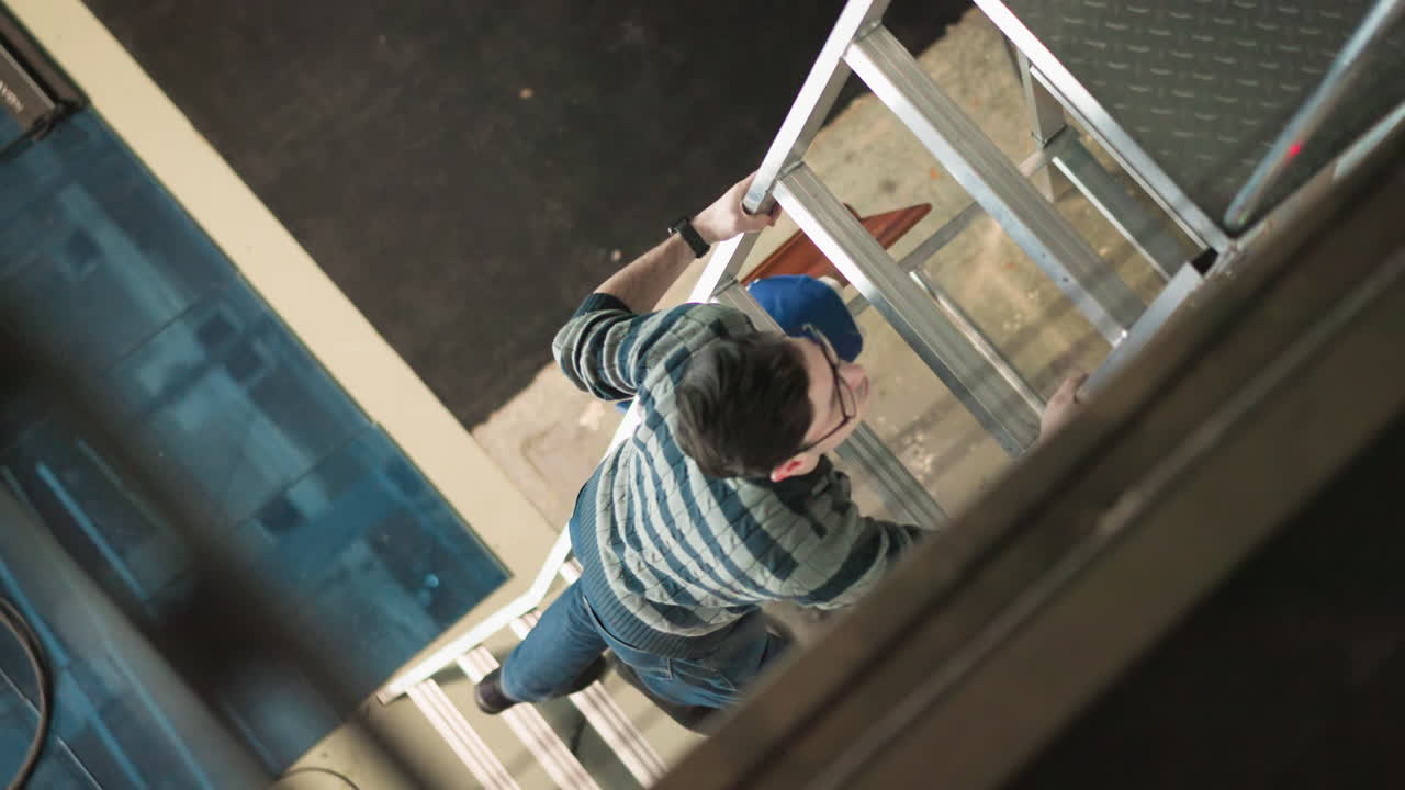 Overhead view shows male technician wearing striped sweater, glasses ascending aluminum ladder inside industrial workspace, gripping rung while performing maintenance amid cables, tiles, surface