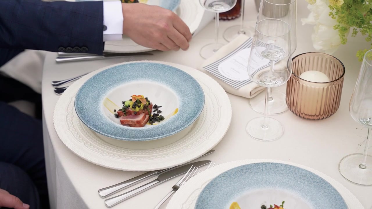 Mani servono antipasti gourmet impiattati su un tavolo elegantemente apparecchiato con piatti dal bordo blu, bicchieri raffinati e una luce naturale soffusa, catturando un'atmosfera da pranzo raffinata ed esclusiva.