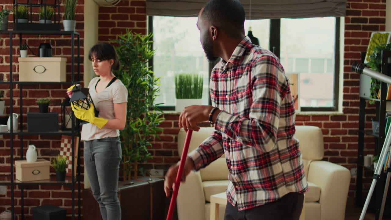 Interracial couple vacuuming floors and cleaning furniture on shelves