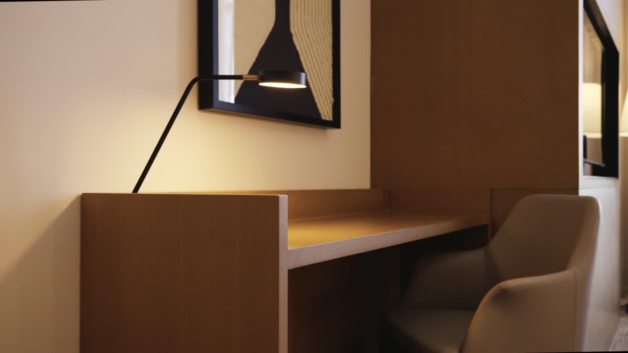Rivelazione con carrello all'indietro da un dettaglio macro in legno a una postazione di lavoro minimalista in hotel con lampada da scrivania di design, sedia ergonomica e arte astratta da parete in un ambiente di ospitalità di lusso.