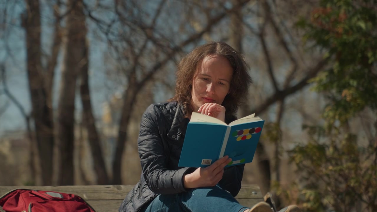 Una donna legge un libro all'aperto su una panchina di legno con uno zaino rosso nelle vicinanze, alberi e cielo sullo sfondo. Lettura all'aperto, parco, luce del giorno, natura, luce solare, primo piano, testurizzato.