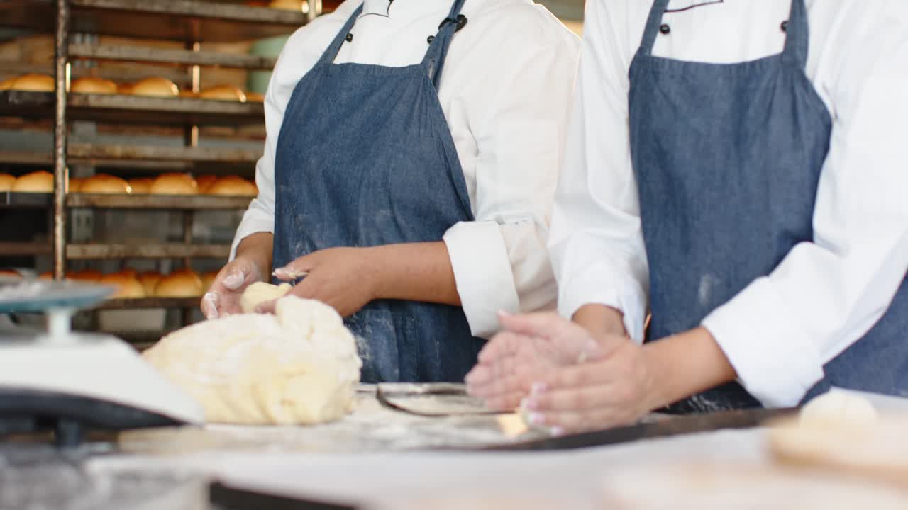 Female bakers in coats forming dough as left baker sprinkling flour by bakery rack for baking