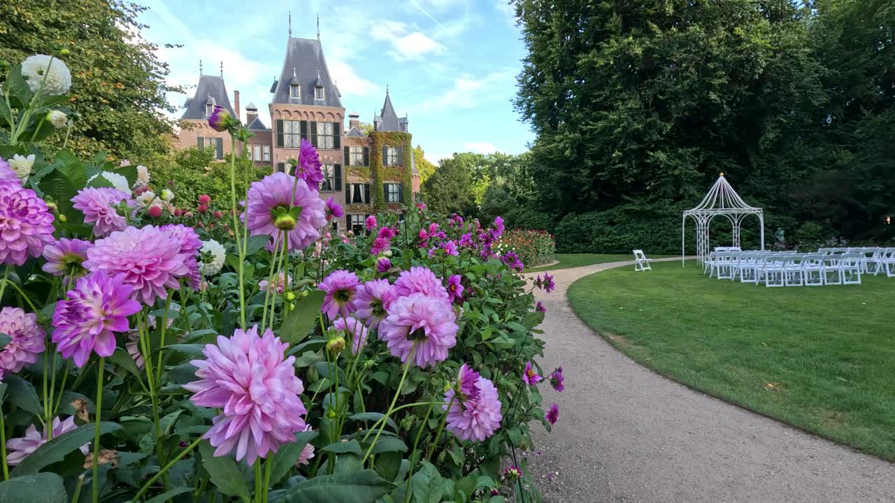 Camera glides past blooming pink dahlias along a winding garden path, revealing a historic castle and white gazebo under bright daylight in Lisse, Netherlands