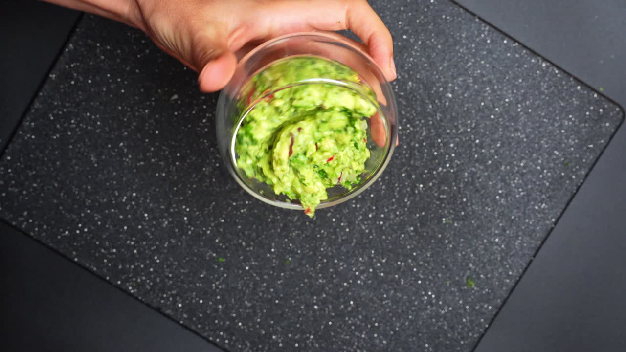 Ruce namáčejí tortilla chips do zeleného guacamole v kuchyni. Ruce sahají pro tortilla chips, aby nabraly guacamole z průhledné misky na černém povrchu.