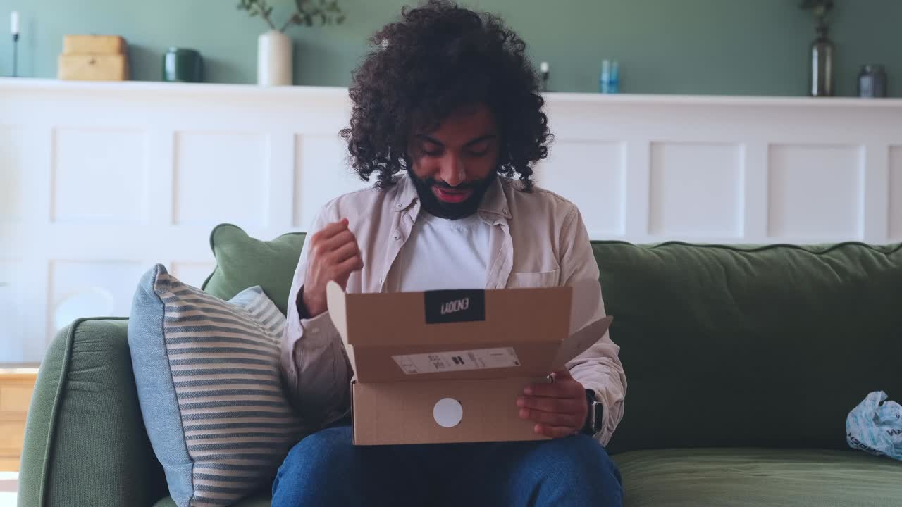 Young arabian man with smile unpack cardboard box sits on sofa in living room
