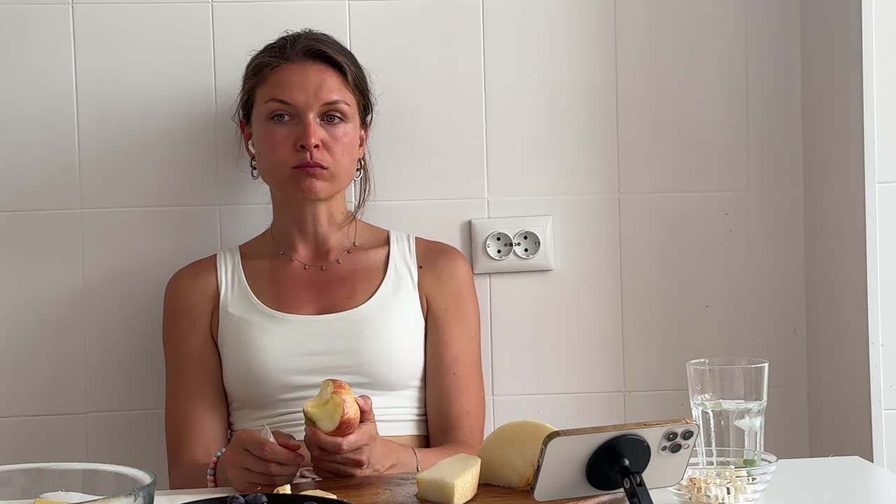 Young Woman Eating an Apple and Watching Her Phone in the Kitchen
