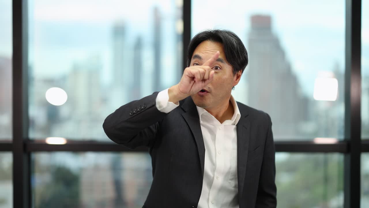 Confident Asian businessman gestures assertively while speaking in bright, modern office with city view