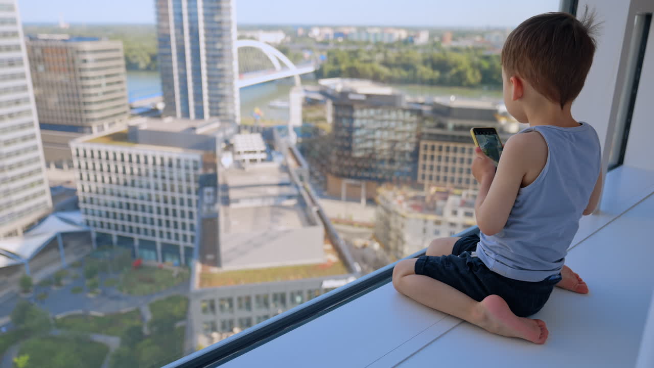Child enjoys view from high-rise window. A young boy sits on a ledge, gazing out at the city skyline and nearby river while holding a smartphone in his hand