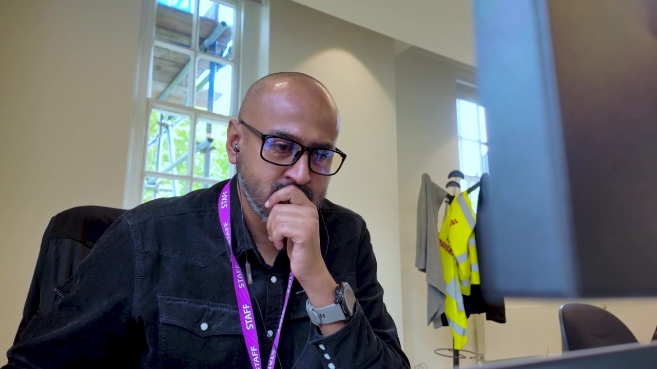 Bald South Asian male wearing glasses and staff lanyard working at an office desk with a computer in front.