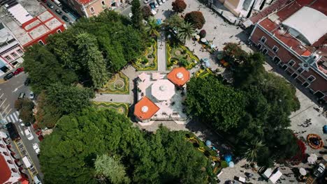 drone moving around the kiosk of mexican town atlixco surround by trees and people passing one evening