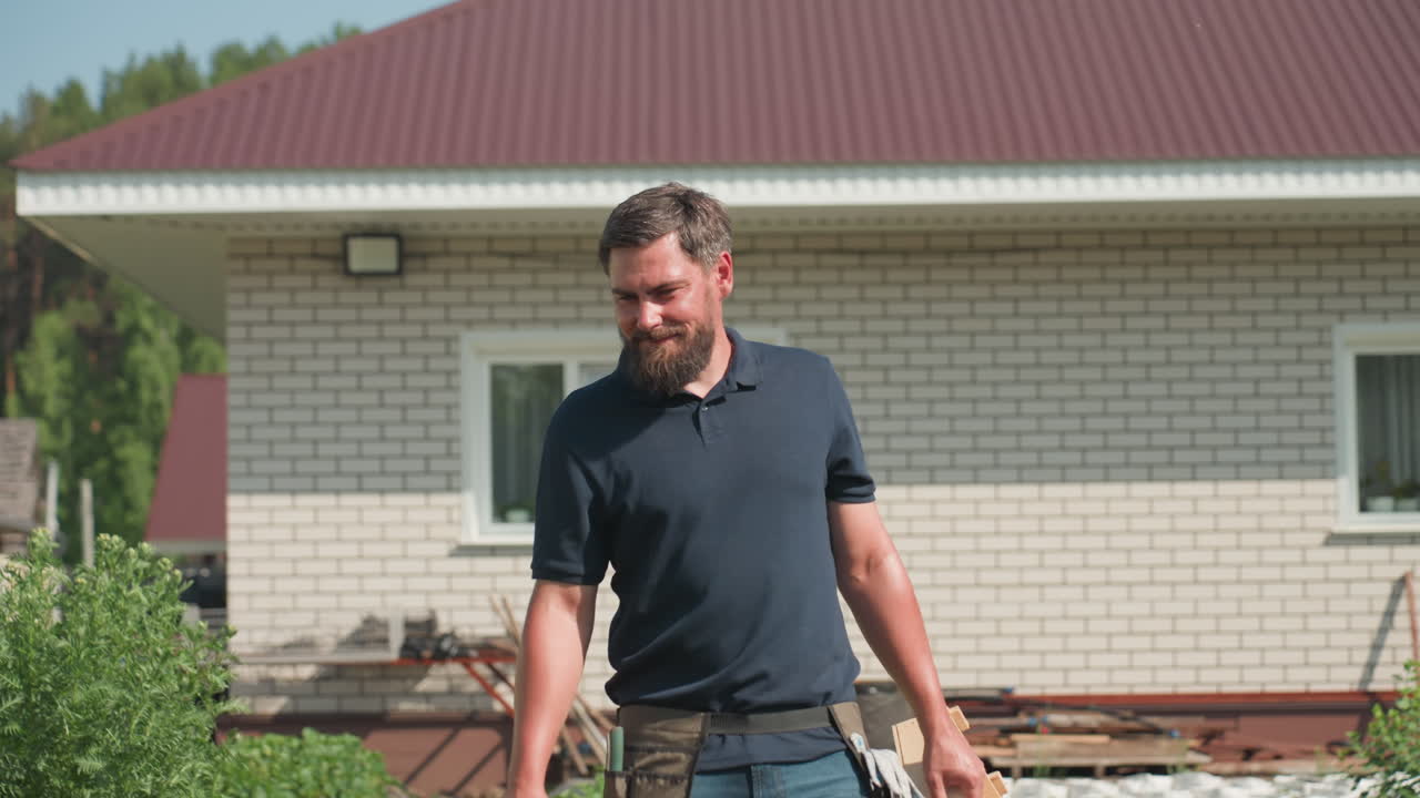 man walking through green crop rows near brick house carrying crate and tools smiling while inspecting farm health and growth on sunny day evaluating plants and enjoying rural work