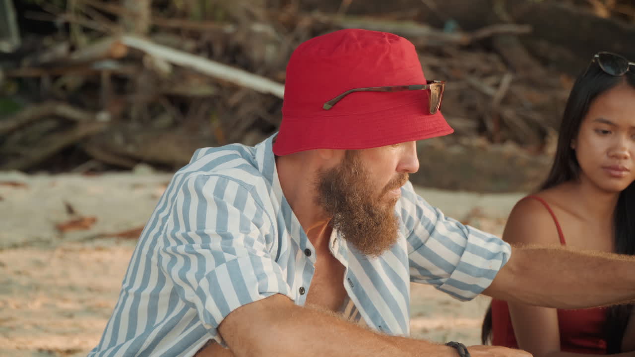 Cerimonia del tè in spiaggia con una coppia, uomo barbuto con cappello rosso versa il tè per una donna in abito rosso sulla battigia sabbiosa vicino a legni, atmosfera rilassata al tramonto, rituale culturale, conversazione intima e sincera
