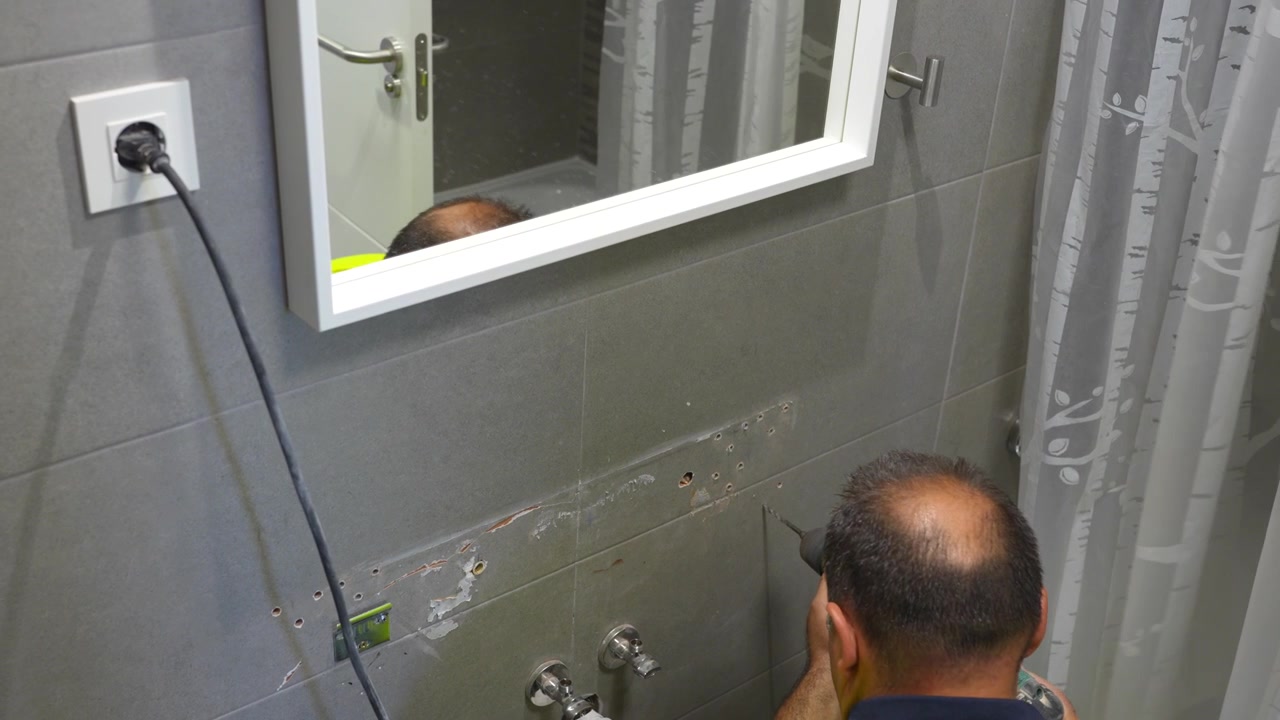 Professional plumber making holes in a tiled bathroom wall using a power drill for renovation