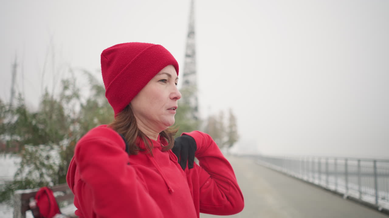 Side view of athlete in red hoodie performing shoulder stretch by turning arm outdoors in snowy park, background features frosted tree, bench, and snow-covered landscape with misty atmosphere
