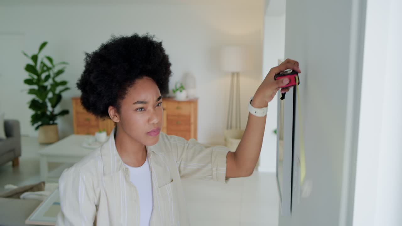 20s woman grabbing tape and level, measuring and hanging frames in living room for even spacing