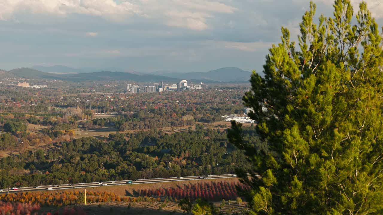Wide view looking towards Woden's high-rise buildings with a highway in the foreground, framed by autumn colours and surrounding landscape.
