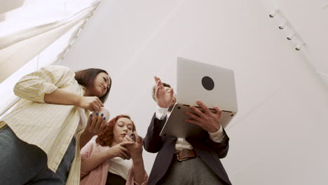 below shot of a man holding a laptop computer and showing something to his female colleagues while standing in the office and talking together 1