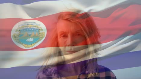 Animation-of-costa-rica-national-flag-waving-over-senior-caucasian-woman-standing-against-sea