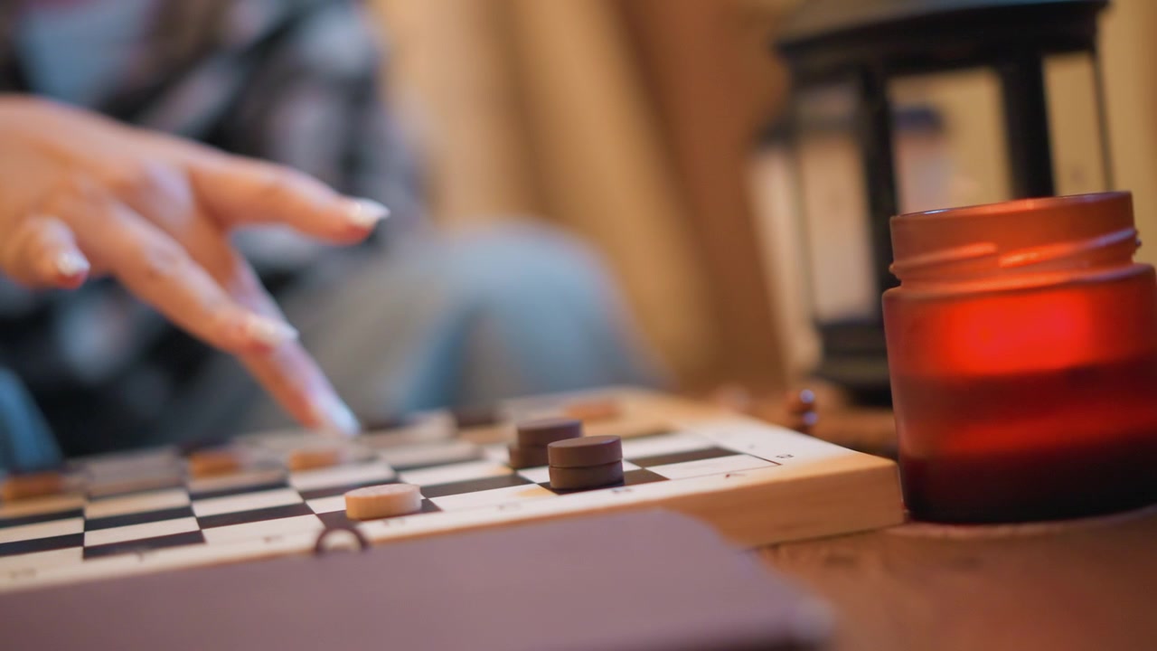 Primo piano della mano di una persona che muove i pezzi su una scacchiera da dama con illuminazione soffusa. Una candela accesa e una lanterna forniscono una calda luce ambientale con lo sfondo leggermente sfocato per enfatizzare il fuoco.