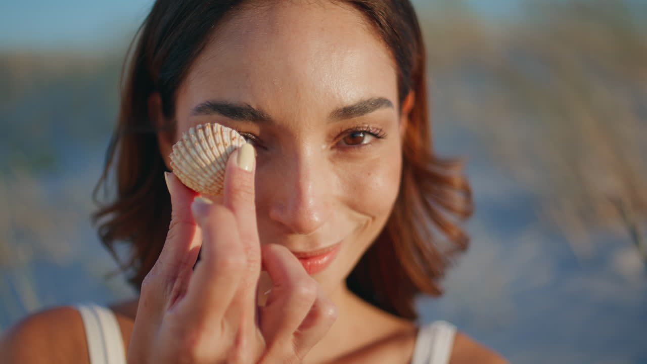 Ritratto di ragazza che si rilassa sulla riva tenendo una conchiglia. Primo piano di donna sorridente con la pelle abbronzata