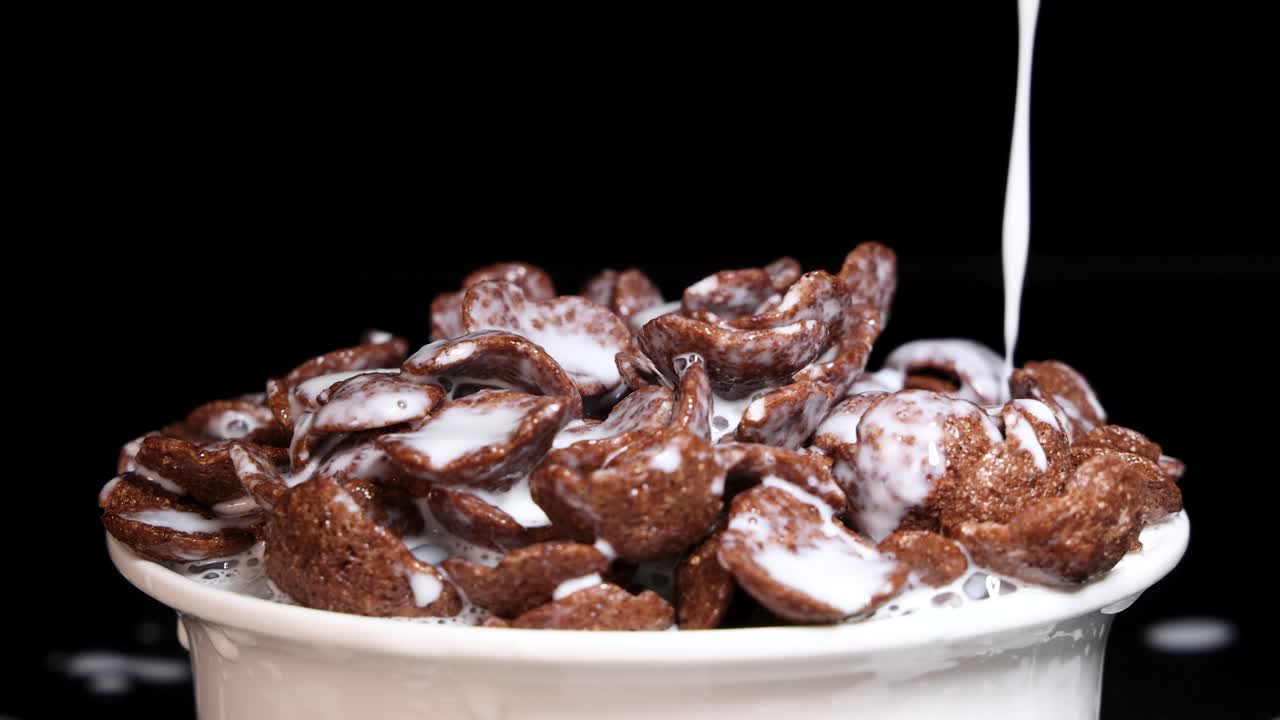 Milk cascades over crunchy chocolate cereal in a white bowl against a dark background, creating a dynamic breakfast scene