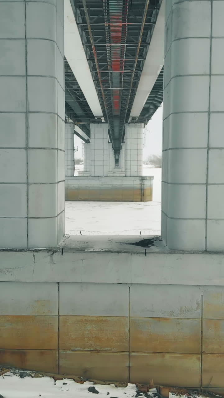 grey bridge support construction in white frozen river against dense forest on bank low angle aerial shot