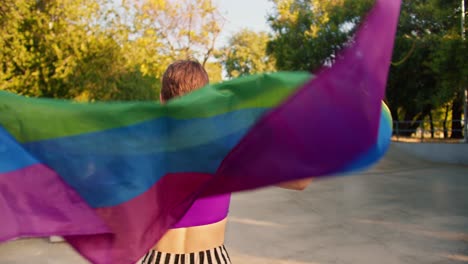 retrato de una chica feliz con una camiseta púrpura y pantalones a rayas que monta patines en un parque de patinaje y sostiene una bandera lgbt que ondea en el viento