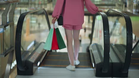 back view of lady in pink dress and short holding three colorful shopping bags and a black handbag, walking onto an escalator as it moves down in a modern mal, in a brightly lit environment