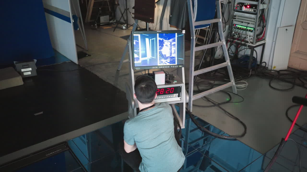 A person working with a touchscreen display in a studio environment, adjusting settings on a control panel while monitoring visuals on a screen. Cables, equipment, and a ladder visible in the background
