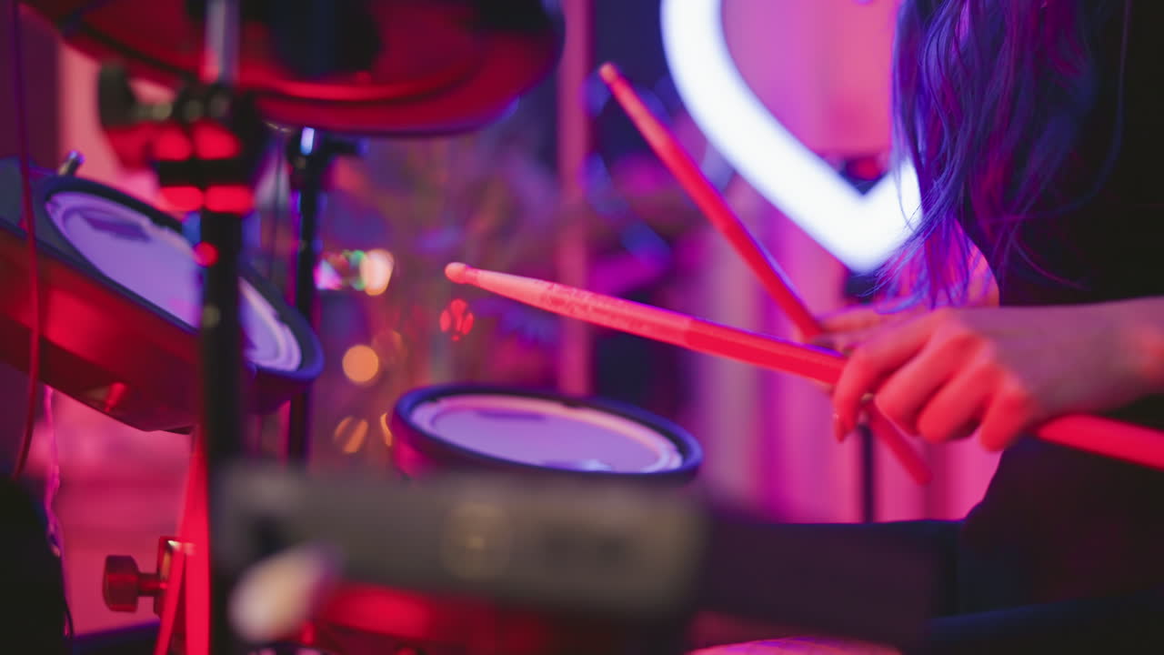 Partial view of lady with long wavy hair covering her face while playing electronic drum set in ambient studio featuring soft neon heart ring light and purple pink background with blurred glow