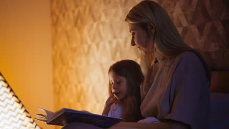 Child-touches-lip-and-reads-book-sitting-with-mom-on-bed