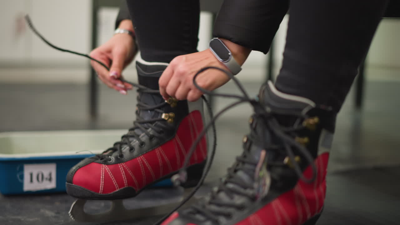 Mulher calçando patins de gelo vermelhos e pretos, amarrando os cadarços com firmeza enquanto se prepara para patinar em local fechado, mostrando foco no calçado com lâminas afiadas refletindo a preparação para a atividade do esporte de inverno e o estilo de vida