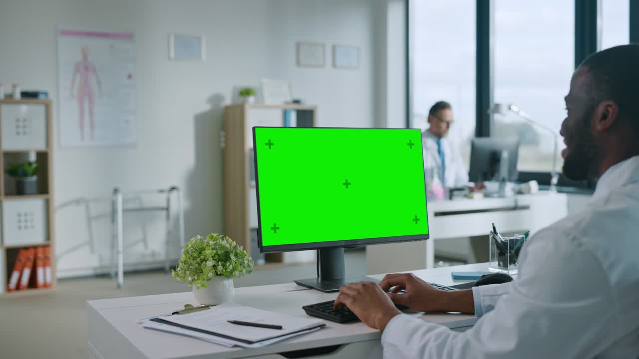 African American Medical Doctor is Working on a Computer with Green Screen Mock Up Display in a Health Clinic. Assistant in White Lab Coat is Reading Medical History Behind a Desk in Hospital Office.