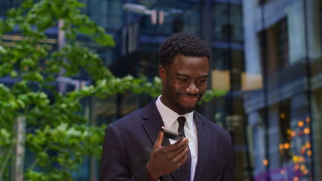 Young-Businessman-Wearing-Suit-Talking-On-Mobile-Phone-Using-Built-In-Microphone-Standing-Outside-Offices-In-The-Financial-District-Of-The-City-Of-London-UK-Shot-In-Real-Time