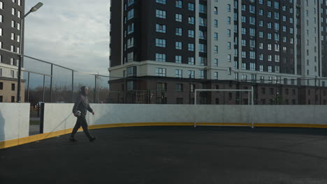 footballer in hoodie and beanie holding ball in right hand steps onto football pitch with goal post in background and tall residential building, dry grass in distant background