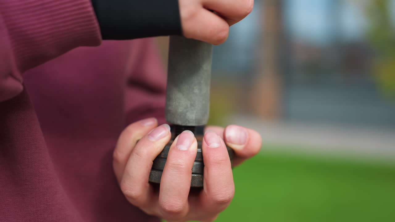 Close up on female fitness coach hands tightening metal dumbbell screw outdoors, fingers secure plates during training prep beside green courtyard lawn, reflecting dedication, precision, progress