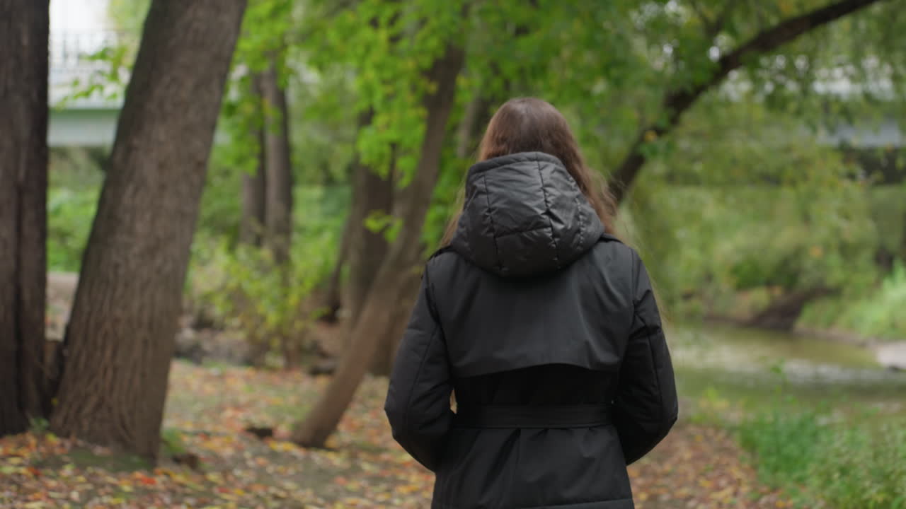 caucasian woman turning walking away from camera backview in hooded jacket strolling along creek path hands in pockets slow steps autumn foliage urban bridge ahead quiet exploration vibe ideal