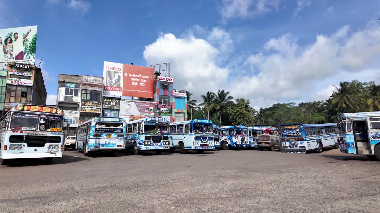 Multiple parked buses at a bus stop garage in Anuradhapura, Sri Lanka, set against a backdrop of colorful buildings and palm trees, under a clear blue sky.