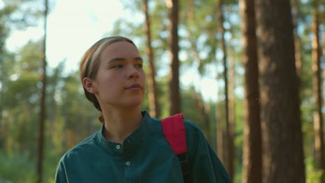 a woman with her hair tied back, wearing green shirt and red backpack, walks through sunlit forest with a warm smile, she gazes thoughtfully around as sunlight reflects gently on her face