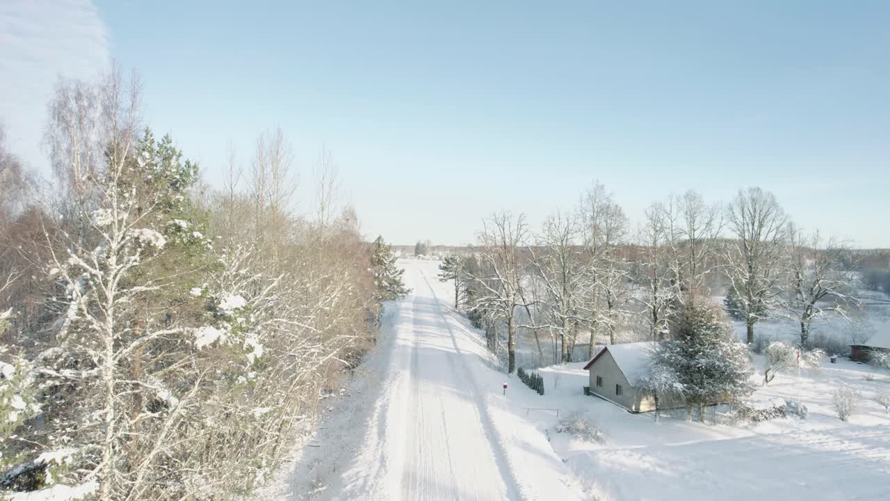 Aerial view of a serene winter countryside featuring snow-covered trees, wooden houses, and a winding road through a frosty forest.
