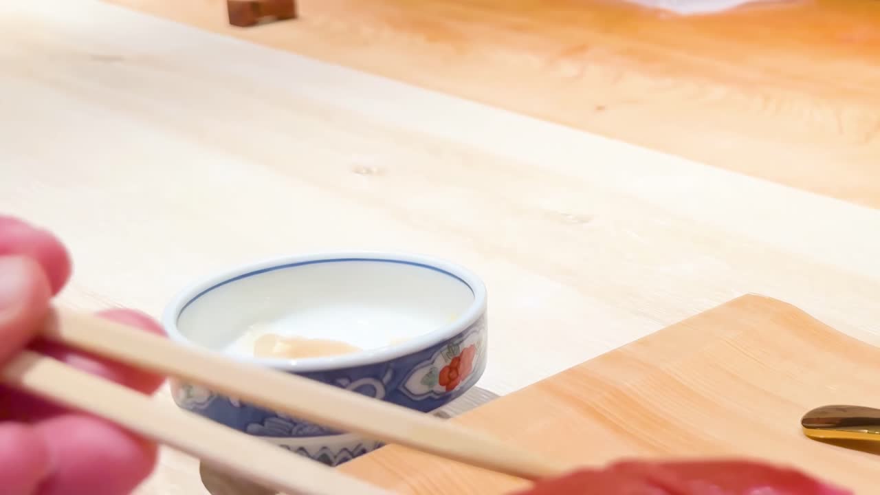 Close-up of chopsticks handling sushi on a wooden board with a small ceramic cup nearby.