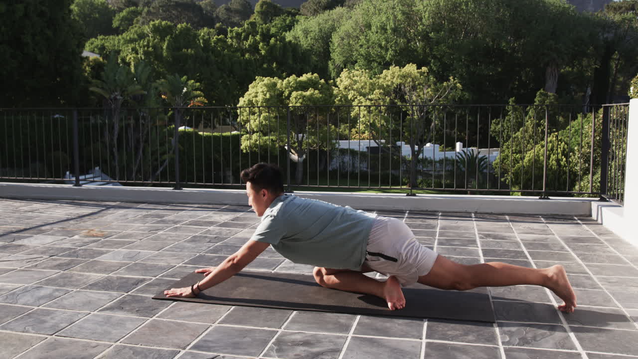 Asian man practicing yoga on patio, stretching on exercise mat outdoors