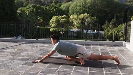 Asian man practicing yoga on patio, stretching on exercise mat outdoors