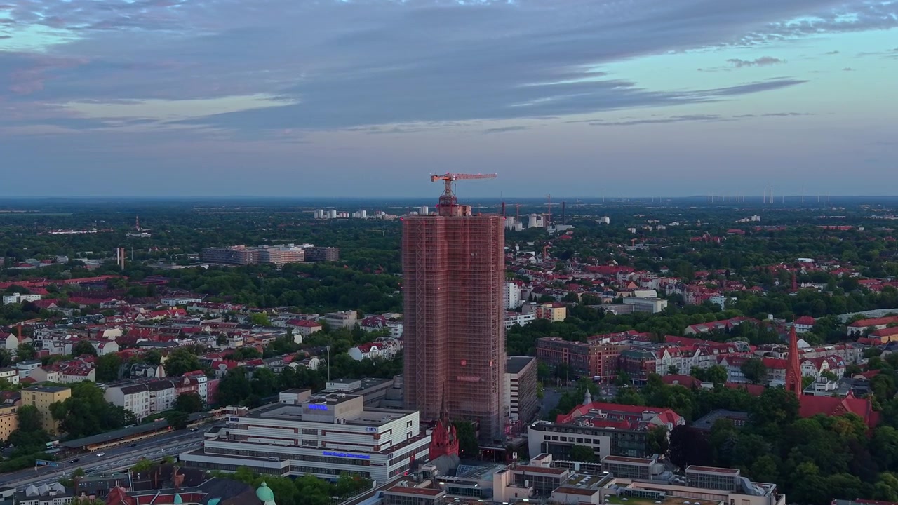 Arranha-céu em construção em uma área urbana com um guindaste no topo, cercado por edifícios residenciais e árvores ao pôr do sol. Panorama aéreo majestoso de drone avançando.