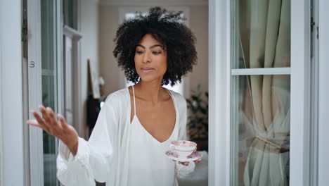 Curly-woman-touching-rain-at-window.-African-lady-looking-camera-holding-coffee