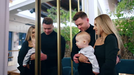 couple with tiny baby boy stand in front of mirrors. father showing the kid's reflection in the mirror.