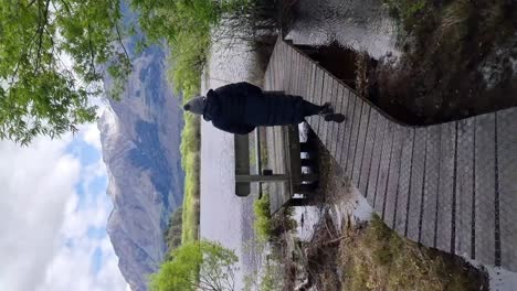 a vertical of a woman walking on a wooden boardwalk with a view of a lake and snow capped mountains in glenorchy new zealand