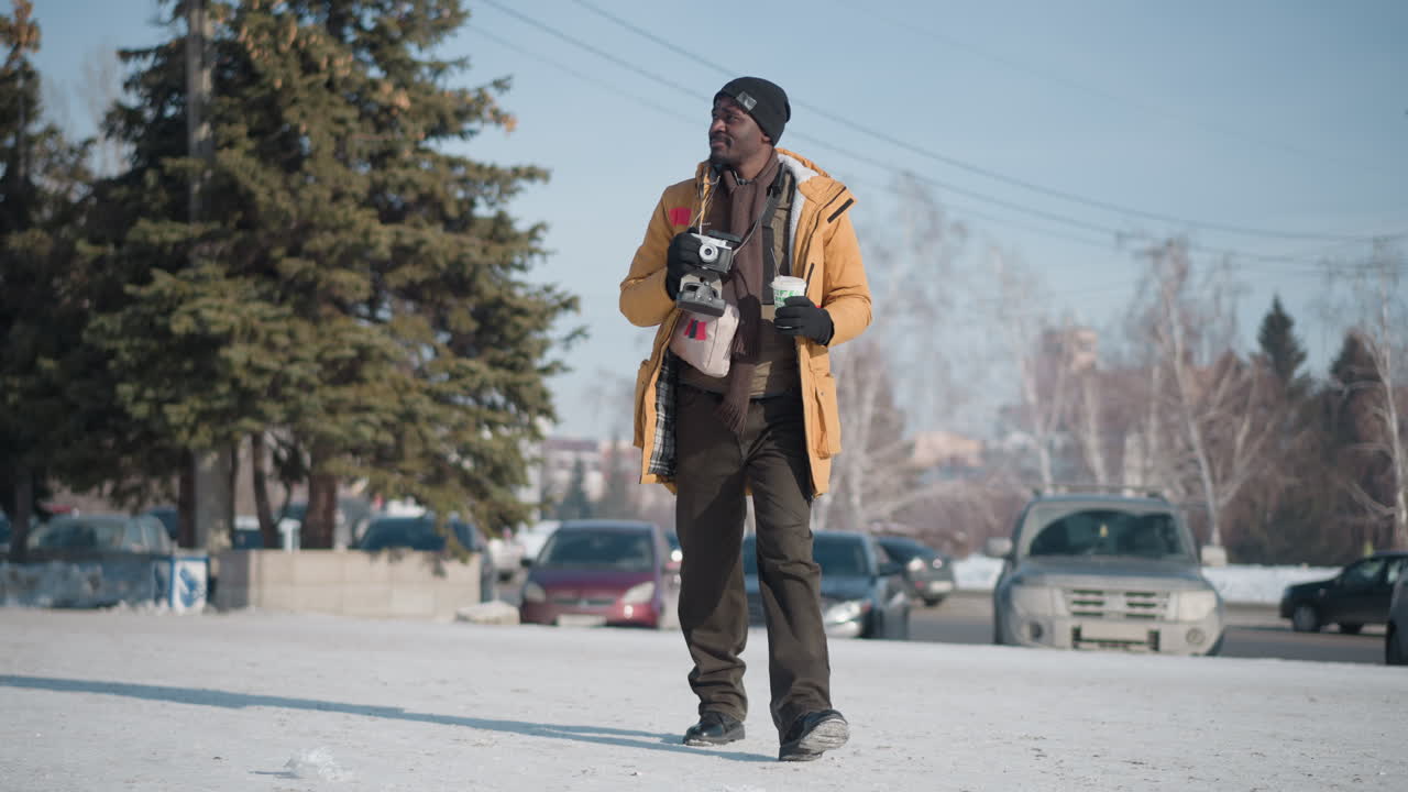 dark man photographer strolling snowy street wearing beanie gloves and winter coat, holding camera and cup of tea, smiling sideways under bright sun as parked cars cast shadows on snow